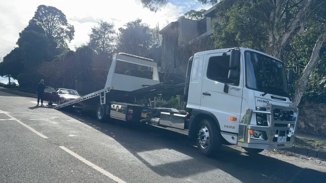 White tow truck parked on a tree-lined residential street, hauling an abandoned vehicle
