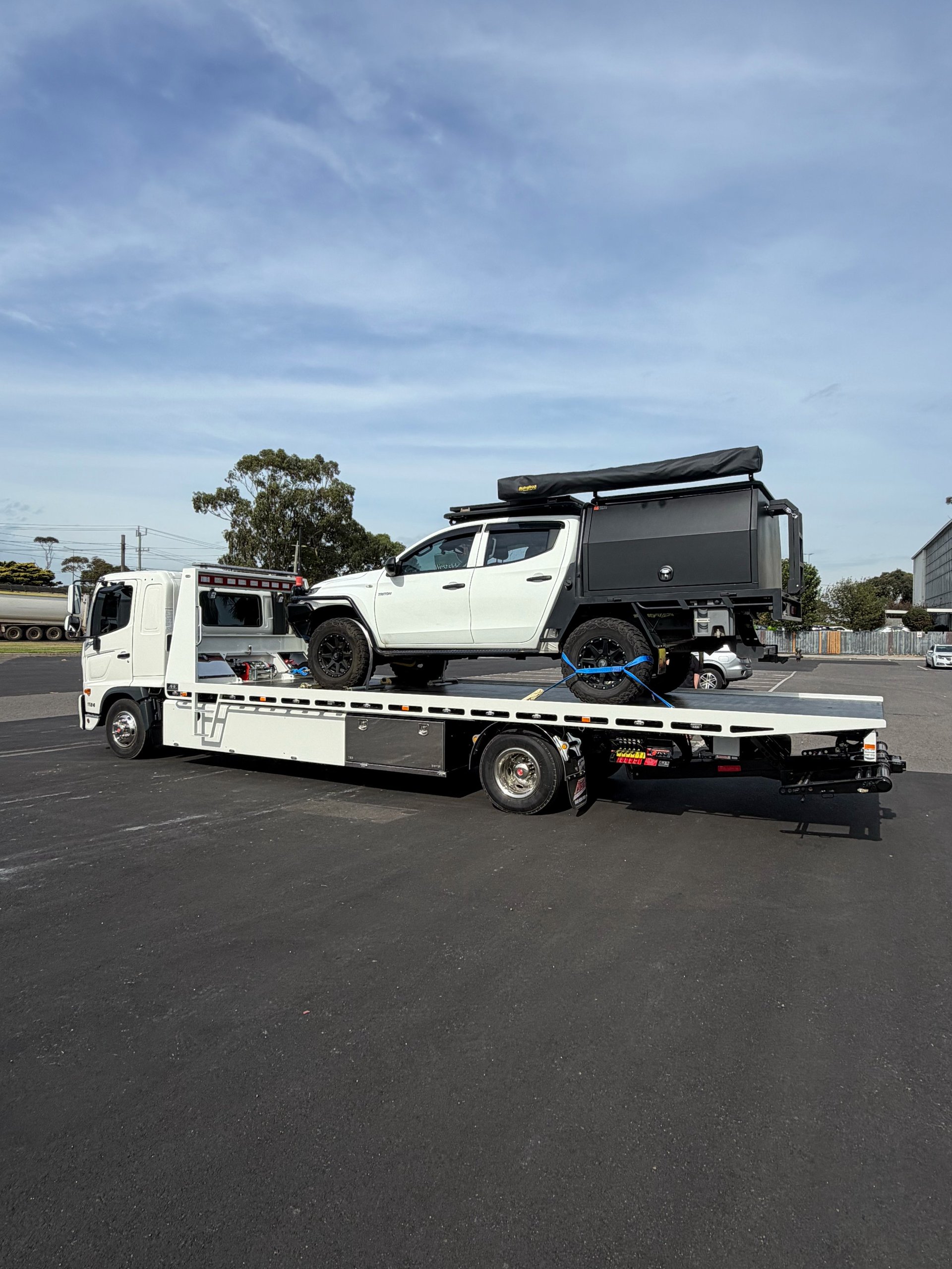 White tow truck carrying a white pickup truck with roof rack on its flatbed in a parking lot