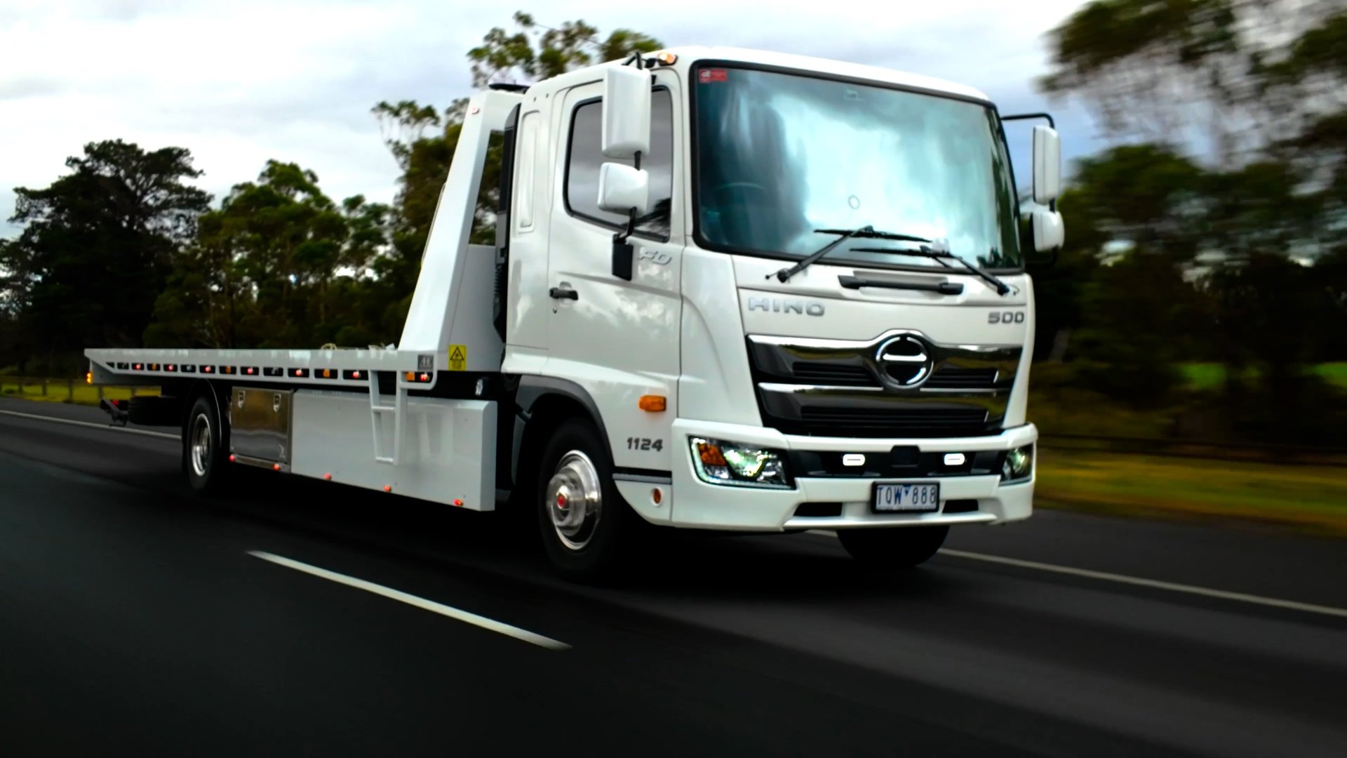 White Hino flatbed tow truck driving on a highway with trees in the background